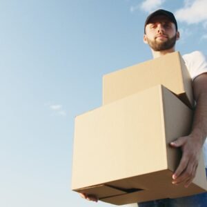 Man in casual attire with beard holding cardboard boxes under a bright blue sky, symbolizing delivery service.